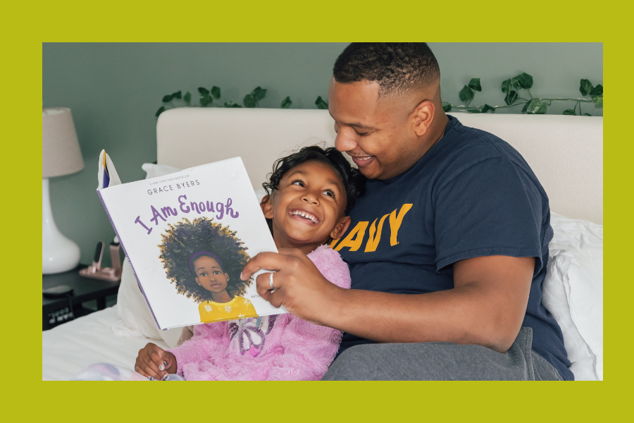 A Black parent and child sit together. They are reading the children's book, I Am Enough. The parent is wearing a Navy branch T-shirt.