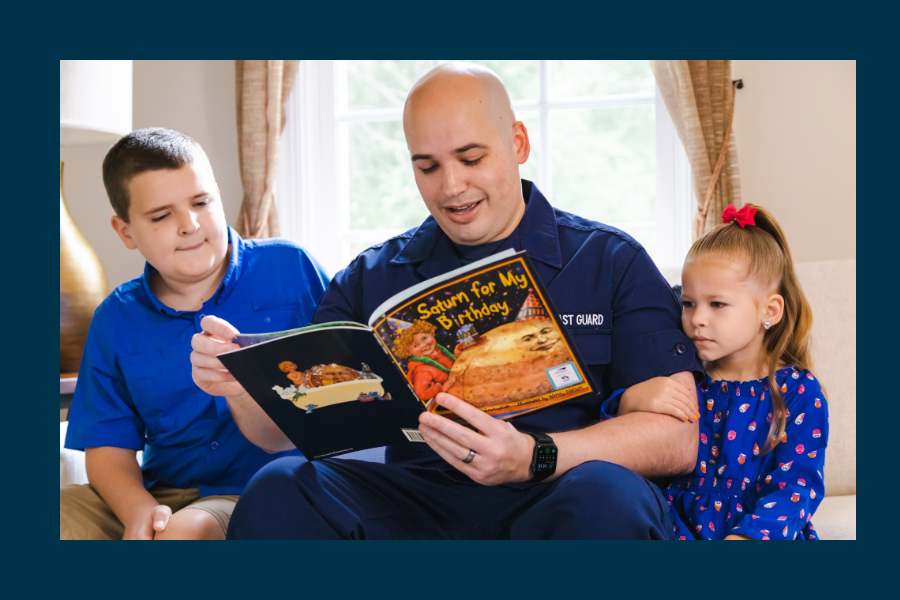 A father in military uniform sits in between his two children. They are reading a children's book together.