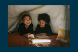Two young children read a book by flashlight underneath a blanket tent.