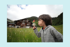 A young child with brown hair and a zip up jacket is in a field. They are blowing on a dandelion. A barn is in the background.