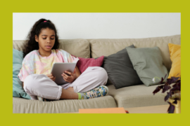 A young girl is seated on a couch. Her legs are criss crossed. She is reading from a tablet.