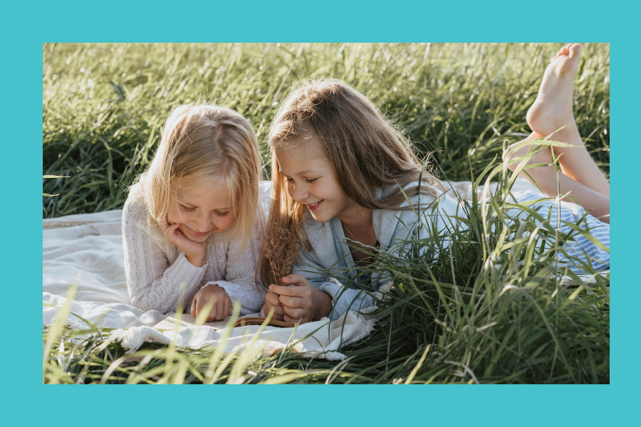 Two young girls are outside in a field. They are laying on a blanket, looking at the same book.