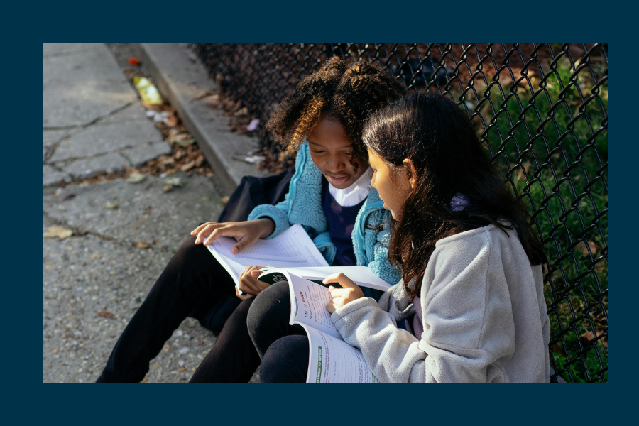 Two young girls are seated on the ground reviewing their homework.