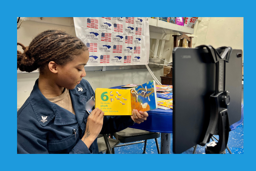 A service member in uniform reads aloud from a children's book titled during a story recording session. The background features a table with books and a United Through Reading banner. A recording device on a tripod captures the session. The scene is framed by a blue border.