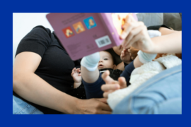 The image shows a close-up of a family reading a book to their baby. The baby, dressed in a dark onesie and wearing white socks, is lying down and looking at the book with curiosity. The parents are partially visible, holding the book and engaging with the child. A soft blanket and a plush toy are also present, adding to the cozy and intimate atmosphere. The image is framed with a blue border.