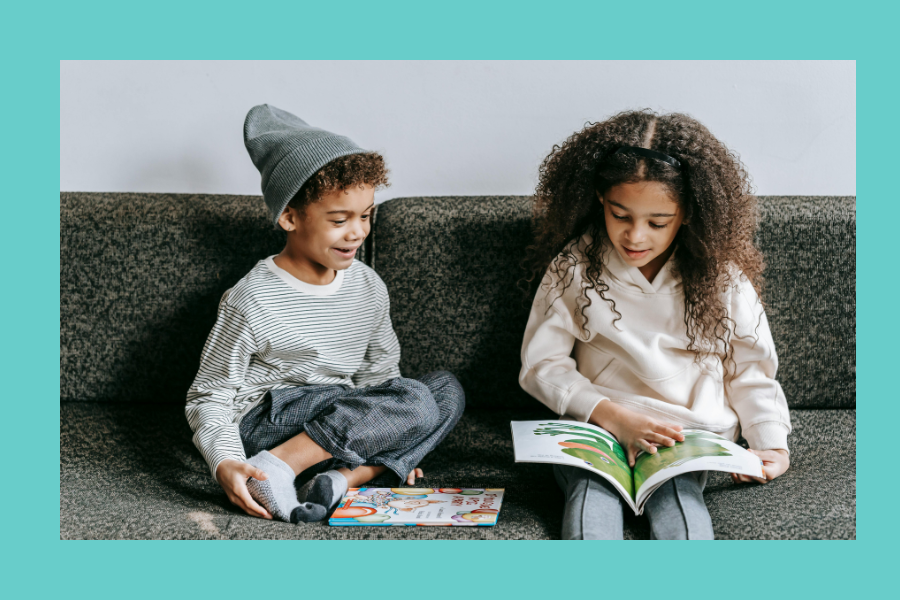 Two young children are sitting on a gray couch reading books. The child on the left, wearing a gray beanie, striped shirt, and gray pants, is smiling and holding a book in their lap. The child on the right, with curly hair and a white hoodie, is fully engaged in reading an open picture book with green illustrations. The background is neutral, and the image is framed by a teal border.