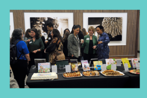 A group of people, mostly women, are gathered around a long black table at an indoor event. The table is set up as a "book tasting," with several children’s books displayed upright on stands, each paired with a food item in front of it. Visible foods include cookies, pastries, and slices of cake. Attendees appear to be engaged in conversation, some smiling and pointing at the display. The atmosphere is warm and social, with modern artwork hanging on the wooden-paneled walls in the background.