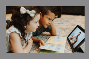 Two young children sit on a carpeted floor, watching a tablet screen that displays a man in military-style clothing reading a children's book. An open picture book with a red whale illustration rests in front of them.
