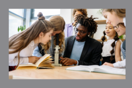 A teacher in a blazer helps a group of young students examine something through a microscope. The children are gathered around a classroom table with open books, smiling and engaged in the STEM activity.