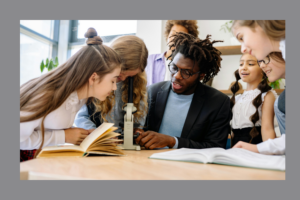 A teacher in a blazer helps a group of young students examine something through a microscope. The children are gathered around a classroom table with open books, smiling and engaged in the STEM activity.