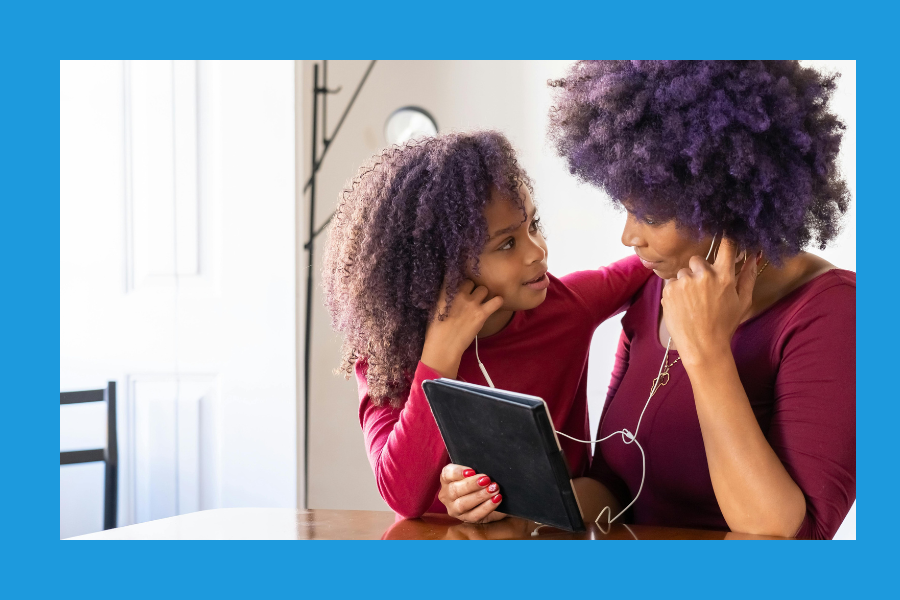 A mother and daughter sit closely together at a table, sharing a pair of earbuds and listening to a tablet. Both wear matching maroon tops and have natural curly hair. The daughter looks up at her mother with a smile as they enjoy a story together. The image is framed with a bright blue border.