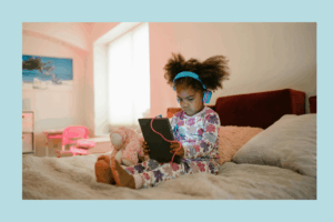 Young girl in floral pajamas sits cross-legged on a bed, wearing bright blue headphones and watching a tablet with a pink charging cable. A pink stuffed animal is beside her, and a soft light from a nearby window brightens the room.