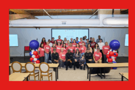 Group photo of about 35 people indoors, gathered in front of a presentation screen displaying “Military Appreciation” with patriotic stars. Many in the front and middle rows wear matching red “Rate” T-shirts, while others wear casual clothing or black shirts with caps. The group stands between two balloon towers made of red, white, and blue balloons. The setting appears to be an event space with wooden beams, large vents, and chairs in the foreground.