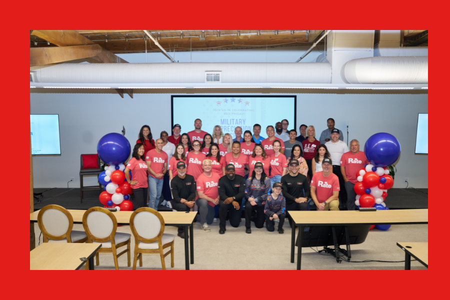 Group photo of about 35 people indoors, gathered in front of a presentation screen displaying “Military Appreciation” with patriotic stars. Many in the front and middle rows wear matching red “Rate” T-shirts, while others wear casual clothing or black shirts with caps. The group stands between two balloon towers made of red, white, and blue balloons. The setting appears to be an event space with wooden beams, large vents, and chairs in the foreground.