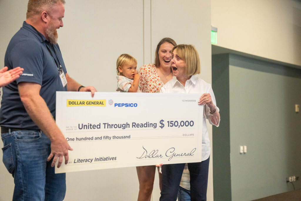 A surprised and joyful United Through Reading representative, Dr. Sally Ann Zoll, receives a $150,000 donation check from Dollar General and PepsiCo, while surrounded by smiling attendees, including Erin Crew and one of her children.