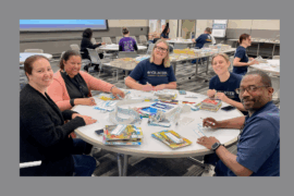 A group of five smiling volunteers sit around a round table, applying labels to children's books. The table is covered with books, stickers, and markers. Other volunteers work at similar tables in the background, surrounded by stacks of books in a large, well-lit room.