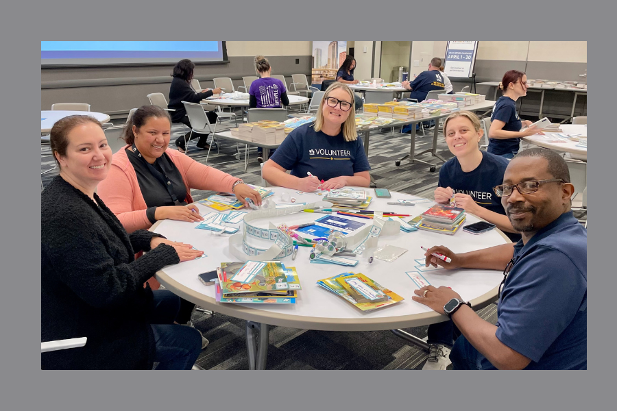 A group of five smiling volunteers sit around a round table, applying labels to children's books. The table is covered with books, stickers, and markers. Other volunteers work at similar tables in the background, surrounded by stacks of books in a large, well-lit room.