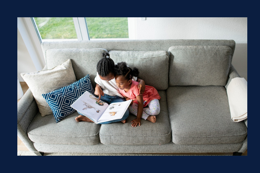 Two young children sit closely together on a gray couch, reading a picture book. One child has their arm around the other as they look at the pages, with patterned pillows beside them and sunlight coming through a nearby window.