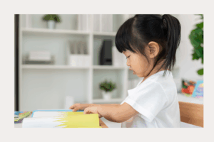 A young child sits at a table, focused on reading an illustrated book with bright colors. The child is turning a page and wearing a white shirt. Behind them is a softly lit room with shelves, plants, and household items in the background.