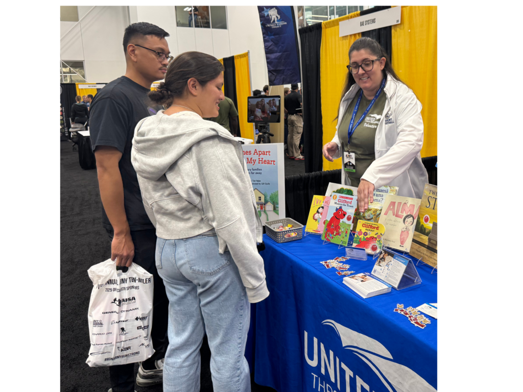 Woman behind a table topped with books. She is speaking to a military couple.