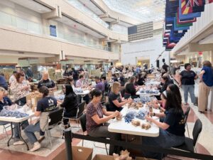 United Through Reading and USAA Volunteers gather in a large indoor atrium to assemble teddy bears and care package items for military families. 