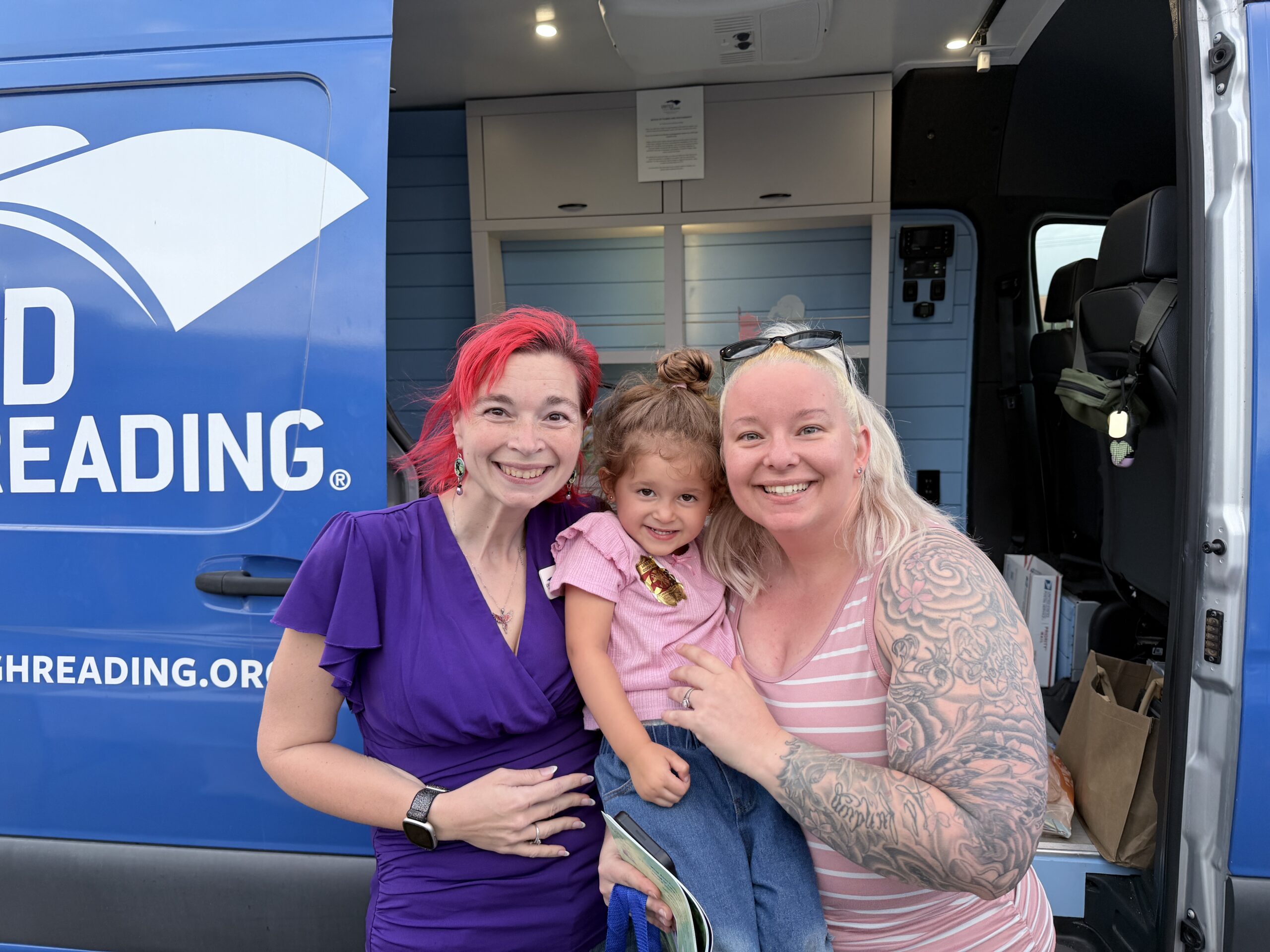 A smiling woman and child pose with a United Through Reading team member in front of a blue mobile reading van.