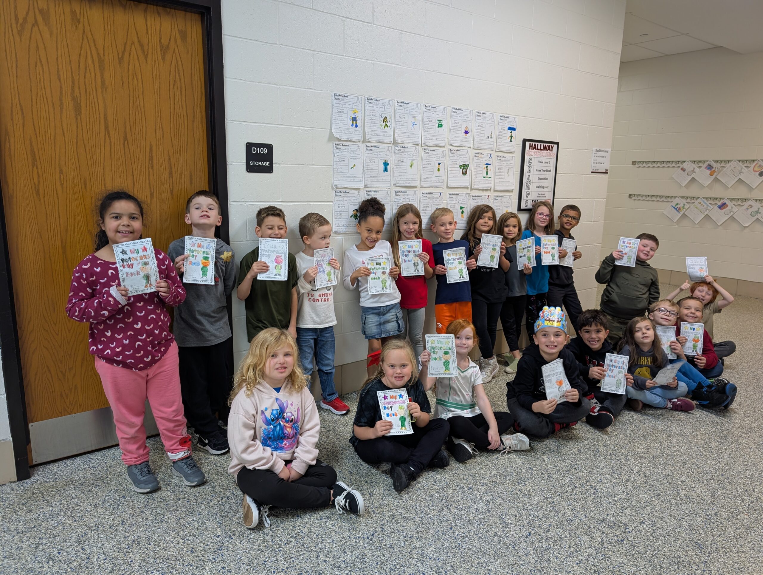 A group of elementary school students stands and sits in a school hallway, smiling and holding up completed reading activity booklets following a United Through Reading literacy activity.