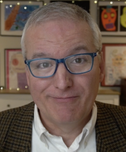 Portrait of Daniel Smith, also known as "The Read Aloud Guy," smiling at the camera. He has short gray hair, wears blue-framed glasses, a white button-down shirt, and a brown plaid blazer. Colorful children's artwork and framed drawings are displayed on the wall behind him, creating a warm, family-friendly setting.