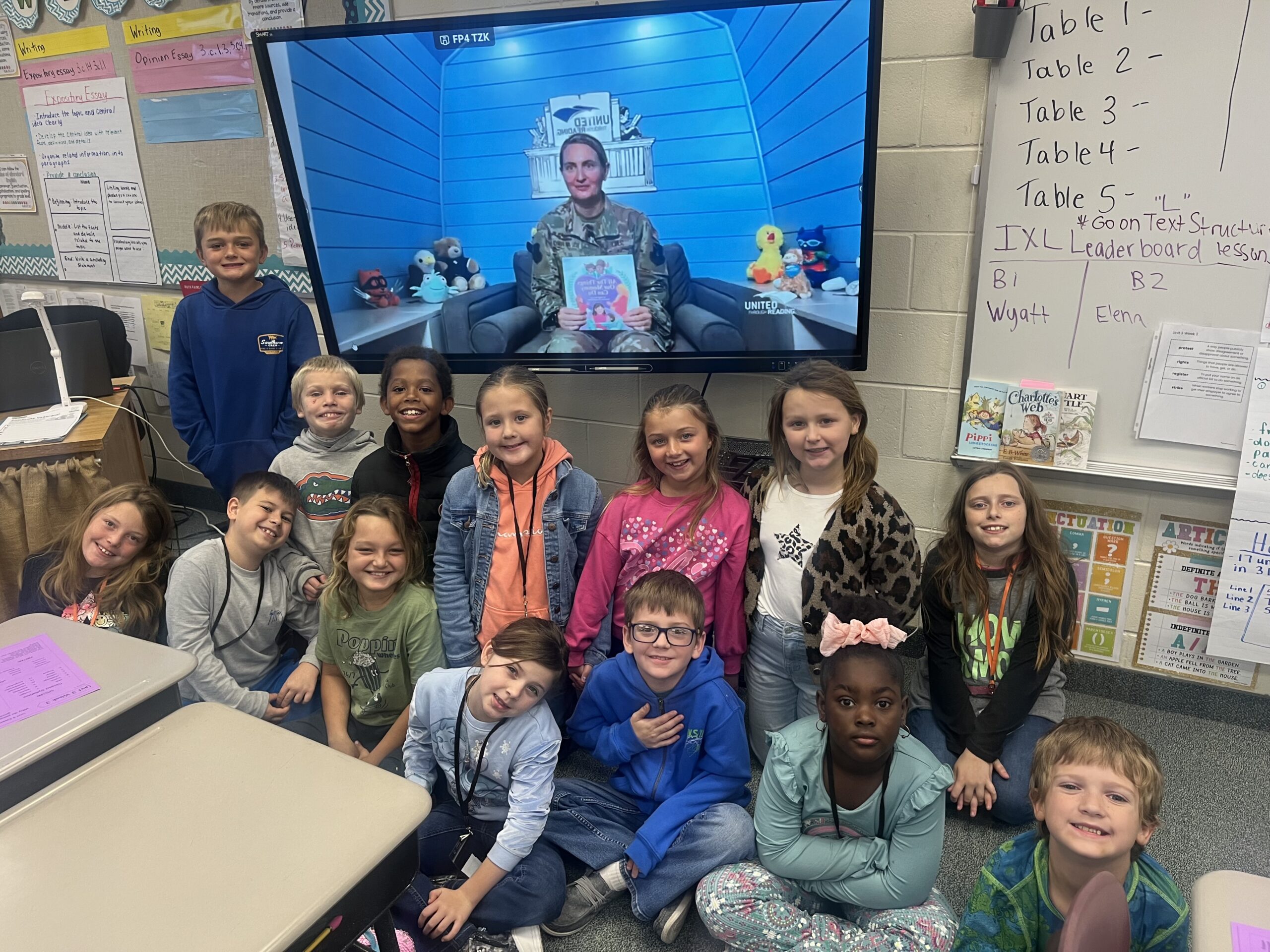 A classroom of elementary school students smiles for a group photo after participating in a virtual United Through Reading storytime. A service member appears on a classroom screen holding up a children's book while reading to the class.