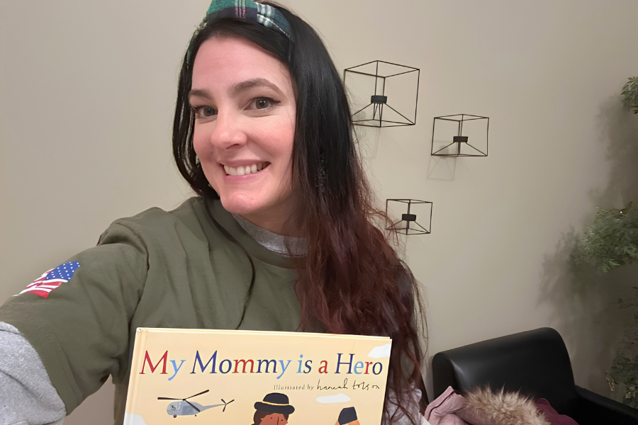 A smiling woman holds up the children’s book My Mommy is a Hero while wearing a military-themed shirt and headband.