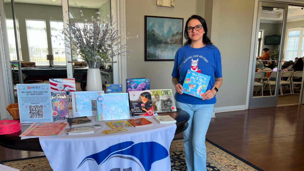 A United Through Reading volunteer holds a children’s book beside a table displaying books and resources at a military community event.