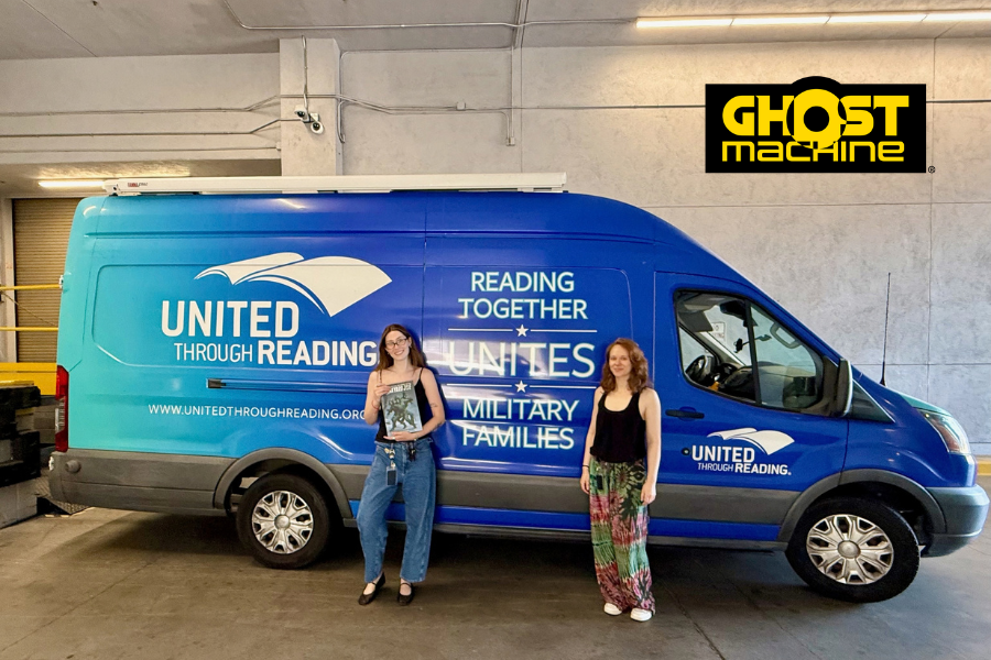 Two Ghost Machine representatives stand beside a United Through Reading Mobile Story Station, with one holding a comic book.