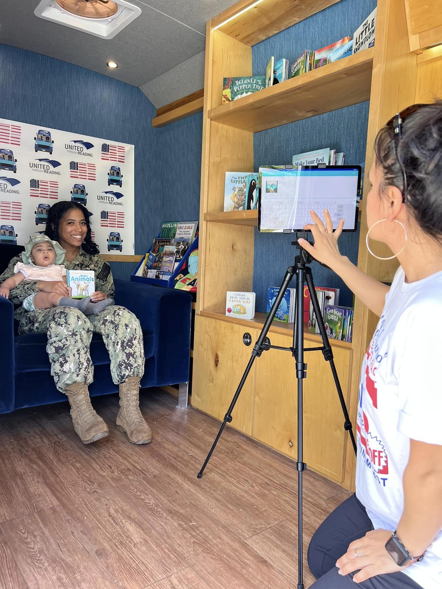 A smiling U.S. Navy service member holds her baby and a children’s book while a volunteer photographs them inside a United Through Reading Mobile Story Station.