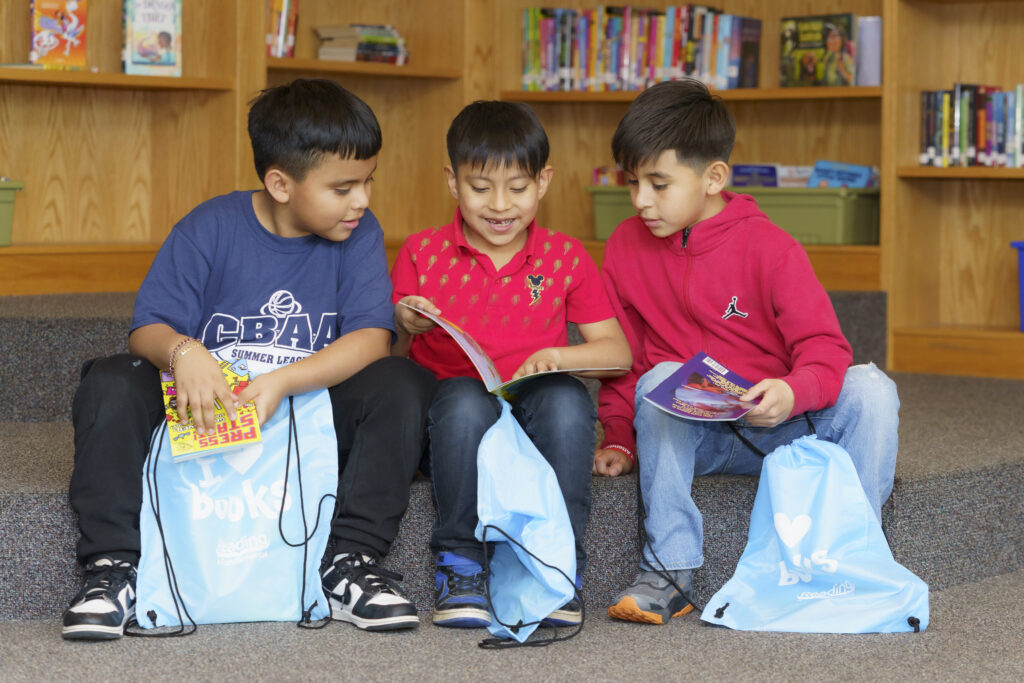 Three children sit together in a library, smiling and reading books beside bags filled with books.