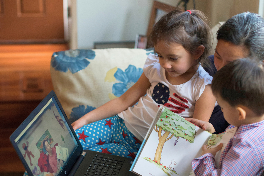 A mother reads with her two children while they watch a loved one’s recorded storytime video on a laptop.