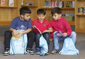 Three children sit together in a library, smiling and reading books beside bags filled with books.