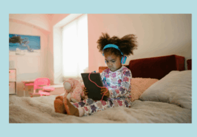 Young girl in floral pajamas sits cross-legged on a bed, wearing bright blue headphones and watching a tablet with a pink charging cable. A pink stuffed animal is beside her, and a soft light from a nearby window brightens the room.