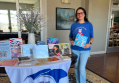 A United Through Reading volunteer holds a children’s book beside a table displaying books and resources at a military community event.