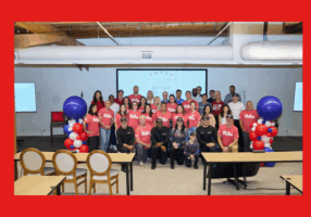 Group photo of about 35 people indoors, gathered in front of a presentation screen displaying “Military Appreciation” with patriotic stars. Many in the front and middle rows wear matching red “Rate” T-shirts, while others wear casual clothing or black shirts with caps. The group stands between two balloon towers made of red, white, and blue balloons. The setting appears to be an event space with wooden beams, large vents, and chairs in the foreground.