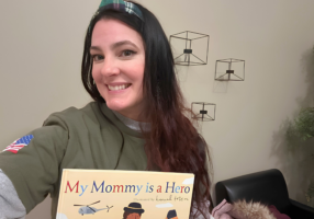 A smiling woman holds up the children’s book My Mommy is a Hero while wearing a military-themed shirt and headband.