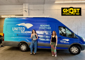 Two Ghost Machine representatives stand beside a United Through Reading Mobile Story Station, with one holding a comic book.