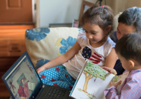 A mother reads with her two children while they watch a loved one’s recorded storytime video on a laptop.