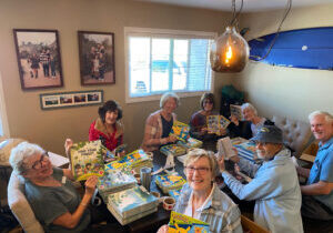 A group of smiling volunteers sit around a table where they are placing stickers on book covers.