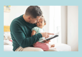 Father reading a book to his toddler daughter while sitting on a bed in a bright room, fostering early literacy and bonding.