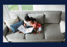 Two young children sit closely together on a gray couch, reading a picture book. One child has their arm around the other as they look at the pages, with patterned pillows beside them and sunlight coming through a nearby window.