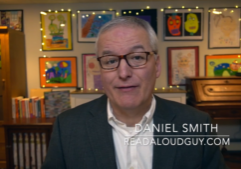 Portrait of Daniel Smith, also known as "The Read Aloud Guy," smiling at the camera. He has short gray hair, wears blue-framed glasses, a white button-down shirt, and a brown plaid blazer. Colorful children's artwork and framed drawings are displayed on the wall behind him, creating a warm, family-friendly setting.