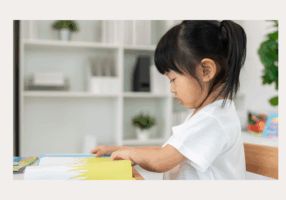 A young child sits at a table, focused on reading an illustrated book with bright colors. The child is turning a page and wearing a white shirt. Behind them is a softly lit room with shelves, plants, and household items in the background.