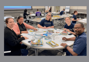 A group of five smiling volunteers sit around a round table, applying labels to children's books. The table is covered with books, stickers, and markers. Other volunteers work at similar tables in the background, surrounded by stacks of books in a large, well-lit room.